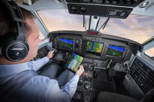 Pilot sitting in cockpit holds iPad up to digital Garmin flight deck. 